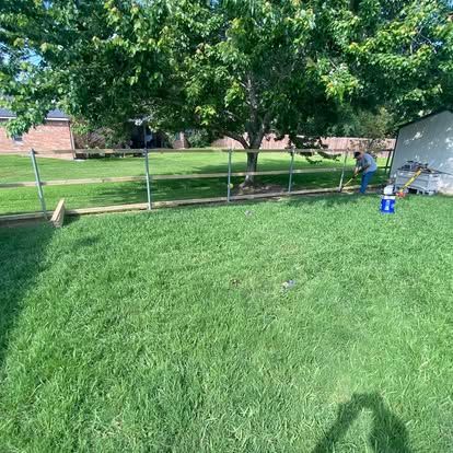 A person installs a wooden rail fence in a grassy backyard near a large tree and a storage shed.