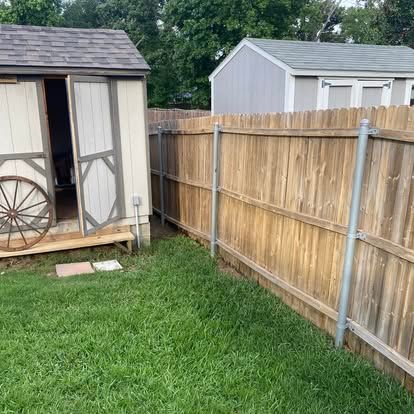 A wooden fence runs alongside a light-colored backyard shed with a wooden wheel leaning against the open door.