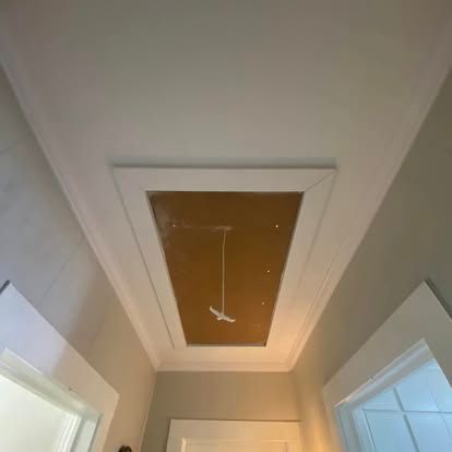 An attic access hatch set into a white ceiling in a hallway, featuring a central pull-down cord.