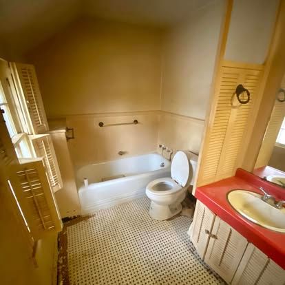 A dated bathroom with a white bathtub, a toilet, a red countertop vanity, and louvered shutters over a window.