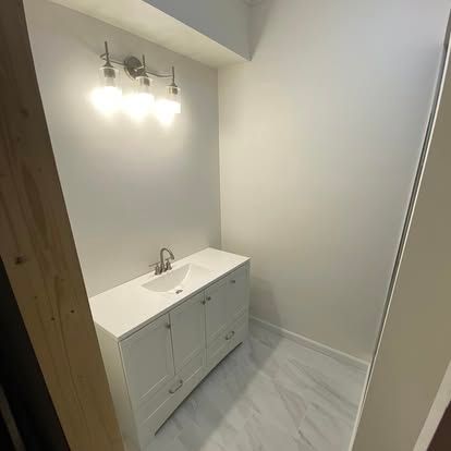 A white bathroom vanity with a chrome faucet, under a three-light vanity fixture in a small room with light gray walls.