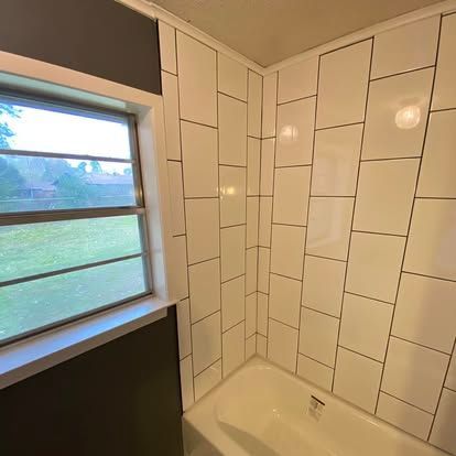 A white tiled shower surround next to a window in a bathroom with dark grey walls.