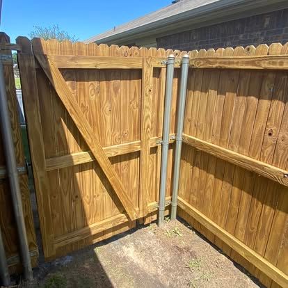 A wooden fence gate with a diagonal brace, reinforced by two metal poles, against a brick house wall.