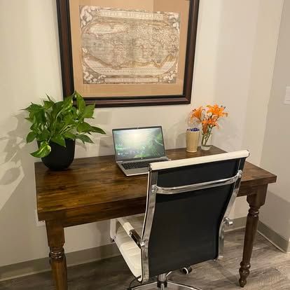 A laptop sits on a wooden desk with a potted plant and flowers, set against a wall with a framed map.