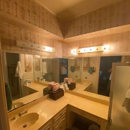 A corner vanity in a bathroom with beige wallpaper, two light fixtures, a sink, and a large L-shaped mirror.
