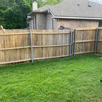 A wooden fence with metal posts borders a grassy yard, with a brick house visible in the background.