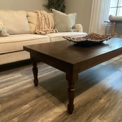 A dark wooden coffee table sits on a wood-look floor in front of a beige sofa with a knit throw and light-colored pillows.