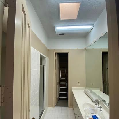 A bathroom with tan walls, a white vanity with a mirror, a glass-enclosed shower, and a doorway leading to a staircase.