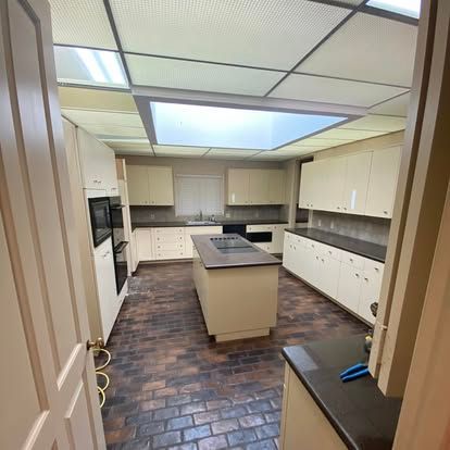 A wide-angle view of a kitchen with light-colored cabinets, a central island, dark countertops, and a brick-pattern floor.