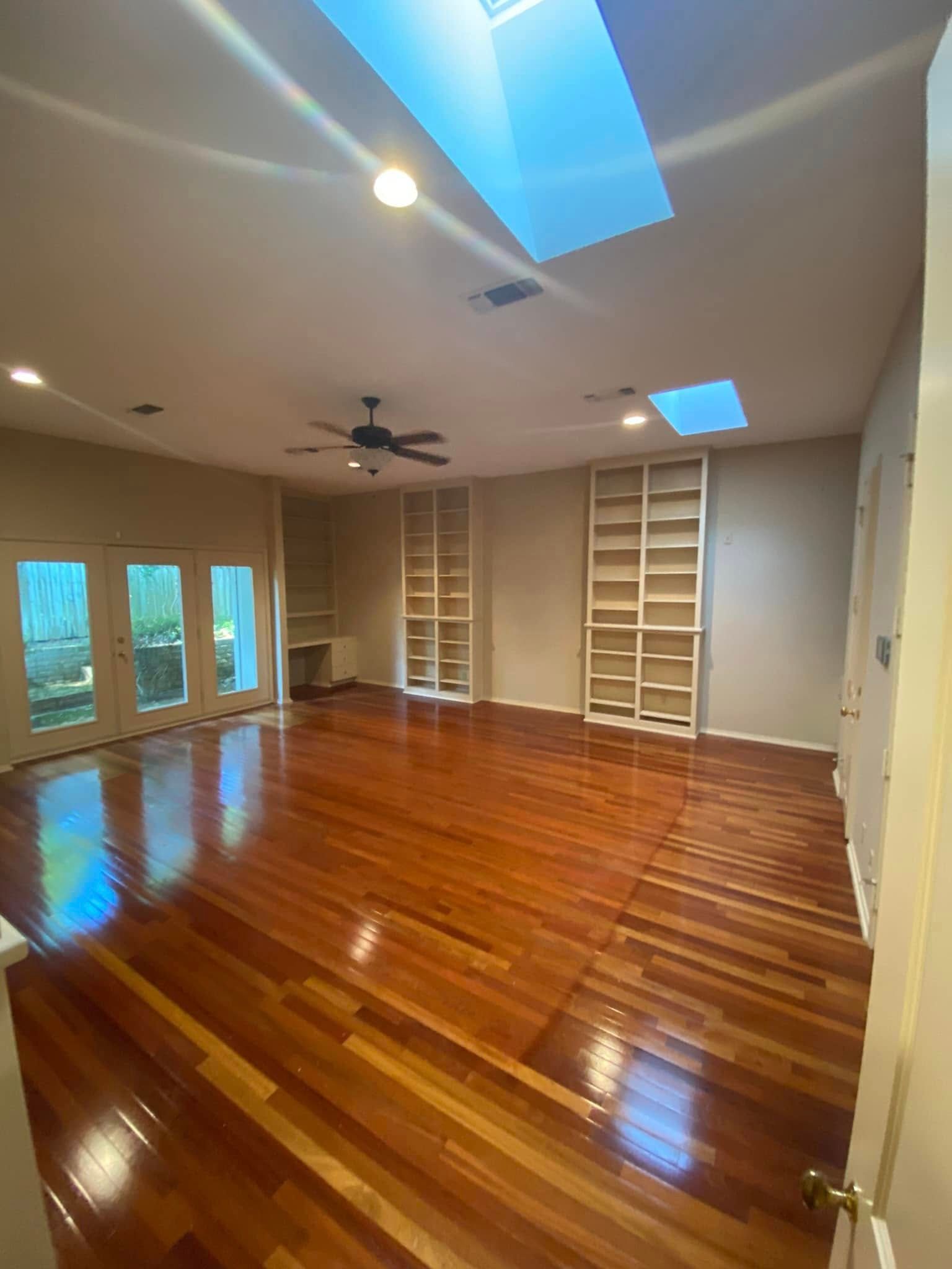 An empty room with polished hardwood floors, two built-in bookshelves, a ceiling fan, and two skylights.
