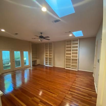 A spacious, sunlit living area with polished hardwood floors, two floor-to-ceiling bookshelves, and glass patio doors.