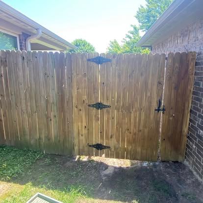 A wooden fence with a gate featuring three black metal hinges and a latch between two brick walls outdoors.