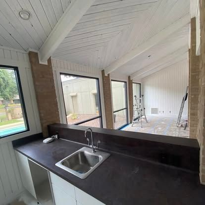 A kitchen counter with a sink overlooks an adjacent room with large windows, white paneled ceilings, and a step ladder.