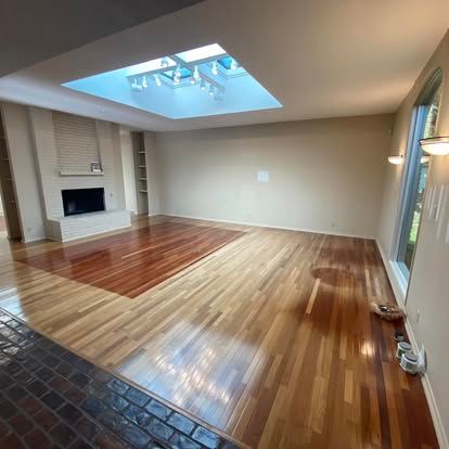 A spacious room with polished hardwood floors, a stone fireplace, recessed lighting, and a large central skylight.
