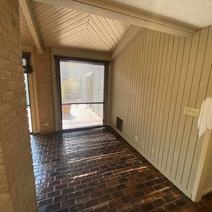 A room with dark brick flooring, vertical light-colored wood paneled walls, a vaulted ceiling, and a large sliding door.