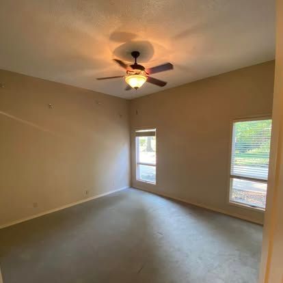A spacious, empty room with beige walls, gray carpet, a ceiling fan with light, and two windows with white blinds.