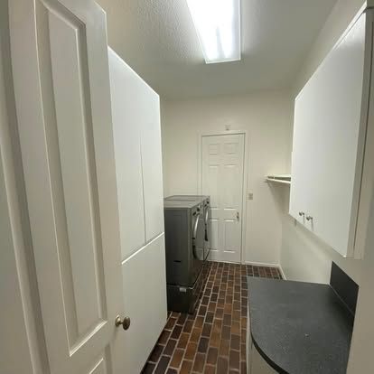 A laundry room with dark brick flooring, a gray dryer, white cabinets, and a doorway at the end of the narrow space.