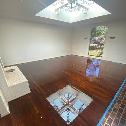 A spacious room with polished dark wood floors, a skylight, a white fireplace base, and a large window looking outdoors.