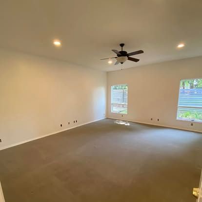 A spacious, empty room with beige carpet, white walls, a ceiling fan, and two windows with view of a fence.