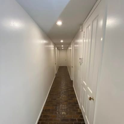 A long, narrow hallway with white walls and a brick floor, illuminated by a row of recessed ceiling lights.