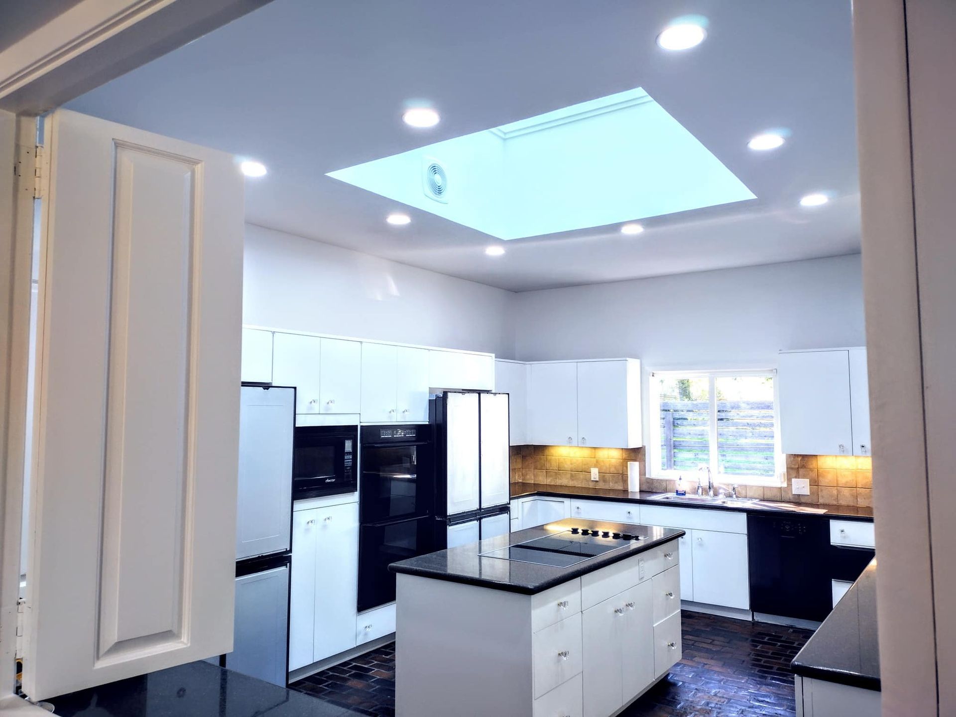 A modern kitchen with white cabinets, dark countertops, a central island, and a large skylight in the ceiling.