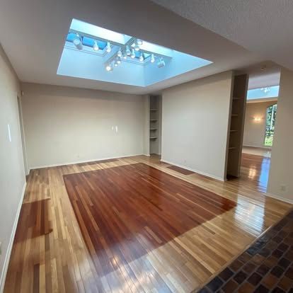 An empty room with a high ceiling, a large central skylight, hardwood flooring, and neutral walls.