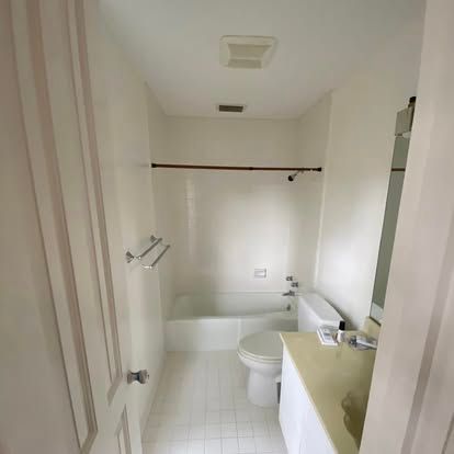 A small, bright bathroom with white walls, white tile flooring, a bathtub-shower combo, and a beige vanity.