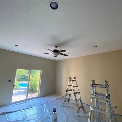 Two metal ladders stand in a room with tan walls and a ceiling fan, with plastic sheets spread across the floor.