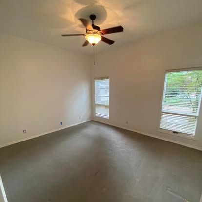 Empty room with beige walls, gray carpeting, a ceiling fan with light, and two windows with closed white blinds.