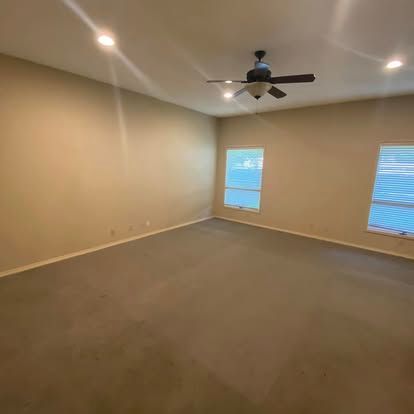 An empty, beige-walled room with gray carpet, a ceiling fan, and two windows allowing natural light to enter.