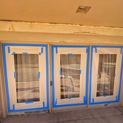 Three sliding glass door panels covered in masking paper and blue painter's tape, set in a beige wall frame.