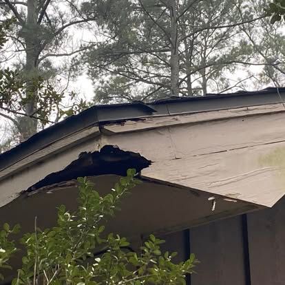 A beige wooden roof eave with a large hole in the fascia board, revealing dark, rotted wood beneath the roofline.