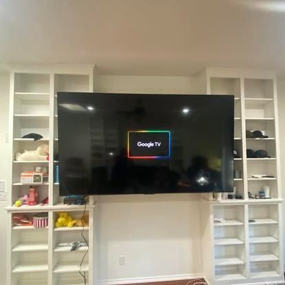 A large flat-screen TV displaying the Google TV logo is wall-mounted between two white, floor-to-ceiling built-in bookcases.
