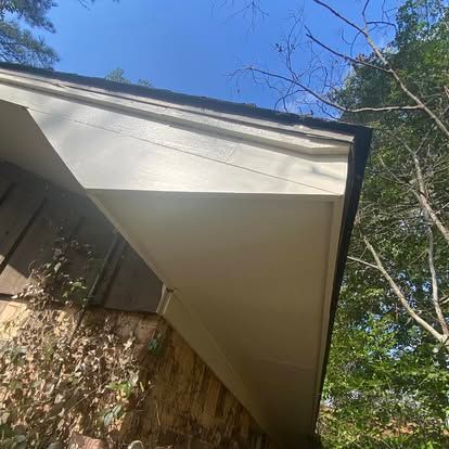 Looking up at the underside of a house roof eave with white trim, next to a textured brown wall and trees against blue sky.