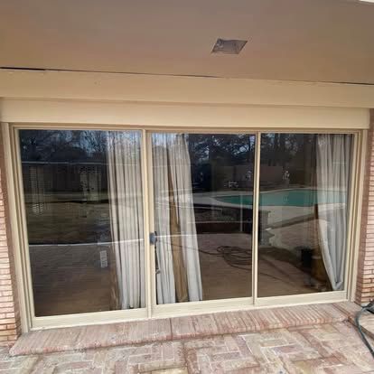 A sliding glass door with beige frames, partially covered by light-colored curtains, overlooks a patio and swimming pool.