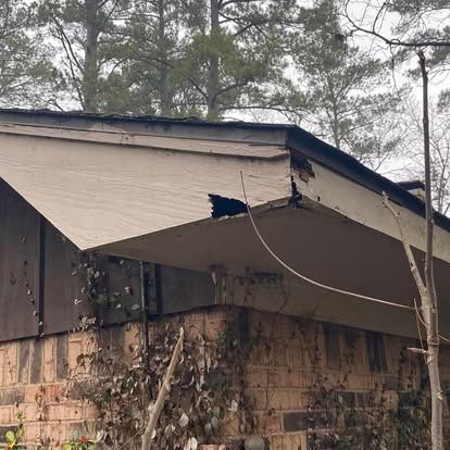 A view of a house roof corner with a large, dark hole in the beige wooden fascia board, indicating rot or structural damage.