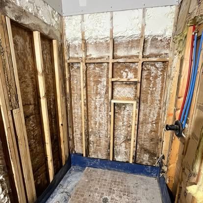 The interior of a shower stall under construction, showing wooden wall studs, insulation, and exposed plumbing pipes.