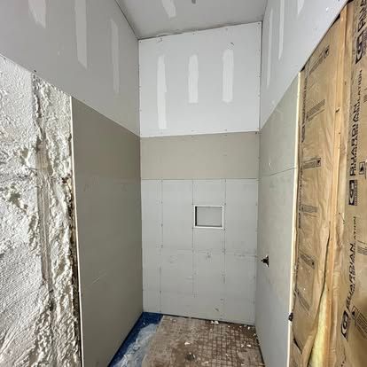 A small, unfinished room under construction, showing grey drywall, exposed insulation, and a tiled shower floor.