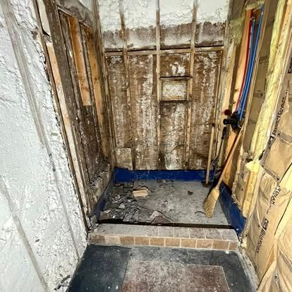 An unfinished shower stall under construction with exposed wooden studs, blue waterproofing, and piping on the right.