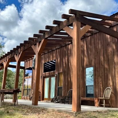 A wooden building with a large, matching timber pergola over a concrete patio under a cloudy blue sky.