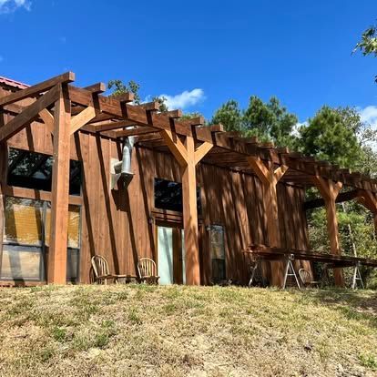 A modern wooden cabin featuring a large timber pergola extension under a clear blue sky, set on a grassy hill.