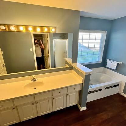 Bathroom with a large vanity, mirror reflecting a person, and a white soaking tub near a window with blue walls.