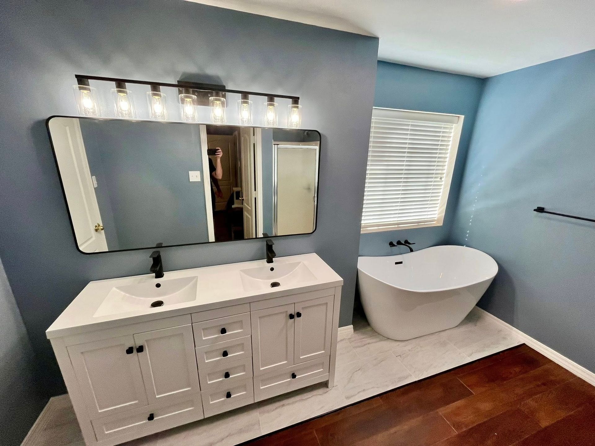 A bathroom with a white double vanity, large mirror with a six-bulb light fixture, and a freestanding white bathtub.