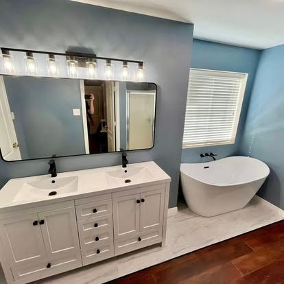 A modern bathroom featuring a white double vanity, a large rectangular mirror, and a standalone soaking tub by a window.