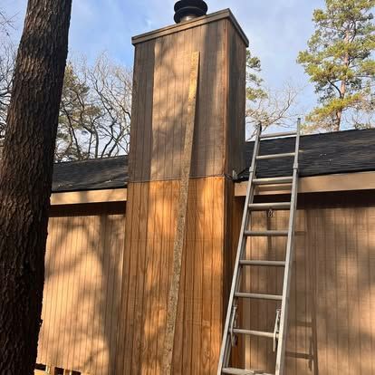 A wooden chimney chase being repaired with new siding panels, next to a ladder against a house roof in a wooded area.