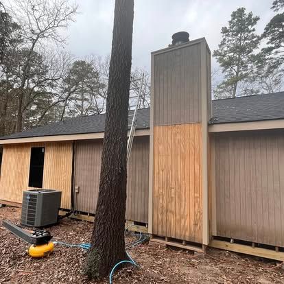 A house exterior features unfinished wooden siding on the chimney and wall, with a tall pine tree and ladder nearby.