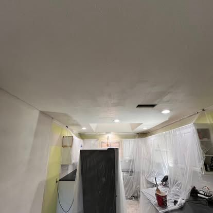 A kitchen ceiling and walls during a home renovation, covered in fresh white paint with plastic sheets draped over cabinets.