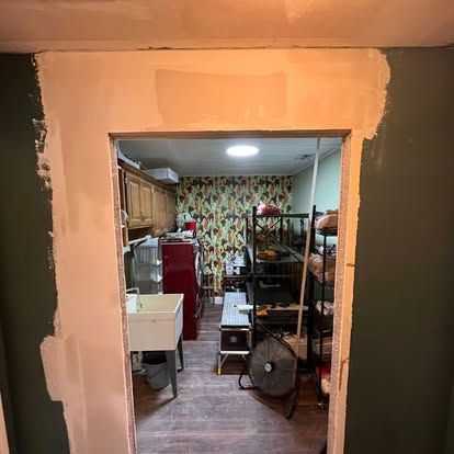A doorway opening with fresh drywall patches surrounding the frame, revealing a room with a utility sink and shelving.
