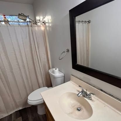 A beige-walled bathroom featuring a vanity with a dark-framed mirror, a toilet, and a light-colored shower curtain.