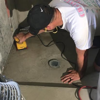 A person in a white t-shirt and black cap uses a yellow sander on a concrete floor near a drain.